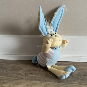 May include: A plush Easter bunny with light blue and cream colors. The bunny has long ears, a blue bow tie, and a striped shirt. The bunny is sitting on a wooden floor.