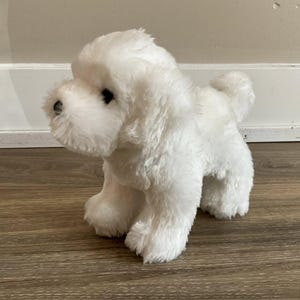Pode incluir: Um cão de pelúcia branco com olhos e nariz pretos. O bicho de pelúcia tem uma aparência fofa e texturizada e está em pé sobre as quatro patas. A cauda do cão está enrolada e sua pelagem é de cor branca brilhante.
