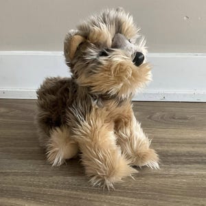 Peut inclure: Une peluche Yorkshire Terrier avec une fourrure marron, beige et grise. L'animal en peluche a un nez noir et de petites oreilles tombantes. Le jouet est posé sur un sol en bois.