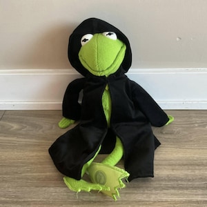 May include: A plush green frog toy wearing a black hooded cloak. The frog has white eyes and a wide smile. The cloak is open in the front, revealing the frog's green body. The frog's feet are visible, with one foot showing some wear.