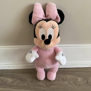 Pu&ograve; includere: Una bambola di peluche di Minnie Mouse in posa eretta. La bambola ha un corpo rosa, un fiocco rosa e orecchie nere. Il suo viso &egrave; color pesca con grandi occhi bianchi, ciglia nere e un naso nero. Indossa guanti bianchi.