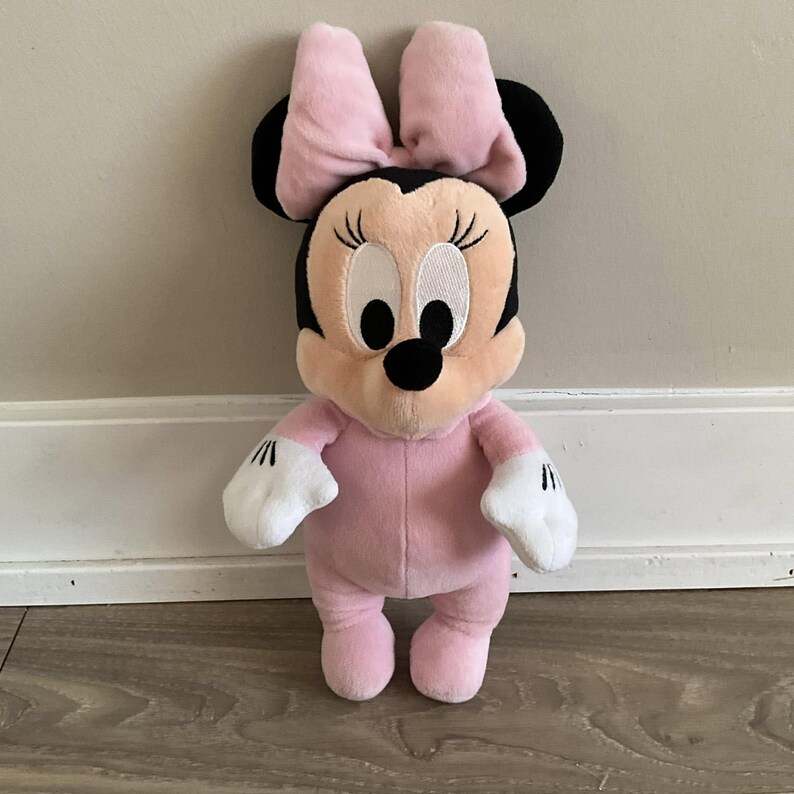 Pu&ograve; includere: Una bambola di peluche di Minnie Mouse in posa eretta. La bambola ha un corpo rosa, un fiocco rosa e orecchie nere. Il suo viso &egrave; color pesca con grandi occhi bianchi, ciglia nere e un naso nero. Indossa guanti bianchi.