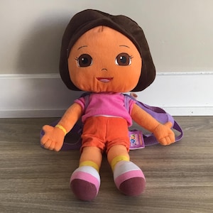May include: A plush Dora the Explorer doll wearing a purple backpack and a pink shirt with orange shorts. The doll has brown hair and a smiling face.
