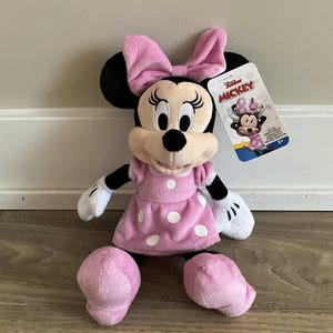 May include: Pink Minnie Mouse plush toy with a pink polka dot dress and a pink bow. The toy is sitting on a wooden floor. The packaging says "Disney Junior Mickey".