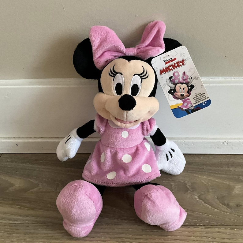 May include: Pink Minnie Mouse plush toy with a pink polka dot dress and a pink bow. The toy is sitting on a wooden floor. The packaging says "Disney Junior Mickey".