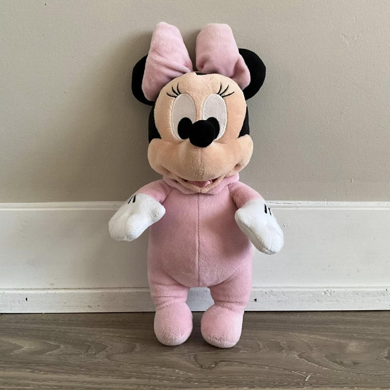 Pu&ograve; includere: Una bambola di peluche di Minnie Mouse in una tutina rosa con un fiocco abbinato. La bambola ha orecchie nere, un naso nero e guanti bianchi. La bambola &egrave; in piedi davanti a un muro bianco.