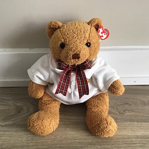 May include: A brown teddy bear wearing a white shirt with a red and black plaid bow tie. The bear has a red Ty Beanie Baby tag attached to its ear.