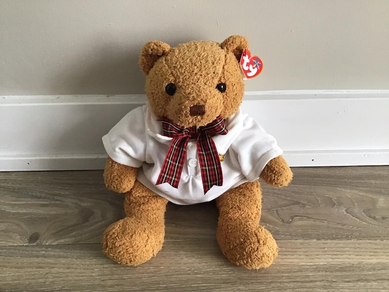 May include: A brown teddy bear wearing a white shirt with a red and black plaid bow tie. The bear has a red Ty Beanie Baby tag attached to its ear.