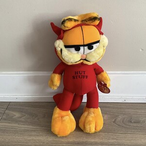 May include: A plush Garfield toy dressed in a red devil costume with horns and a tail. The orange and yellow cat has a grumpy expression and the text "HUT STUFF" on the chest. The toy is holding a small, brown, heart-shaped object.