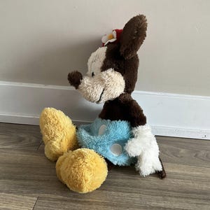 May include: A plush toy of a cartoon mouse with brown and white fur, wearing a red flower in its ear. The mouse has yellow feet, blue shorts, and white gloves. The toy is sitting on a wooden floor.
