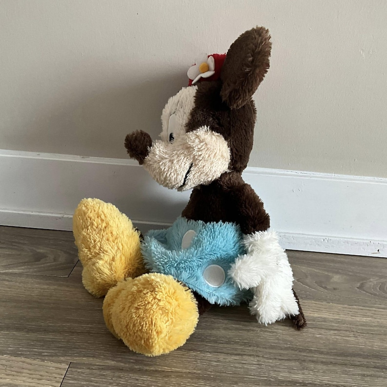 May include: A plush toy of a cartoon mouse with brown and white fur, wearing a red flower in its ear. The mouse has yellow feet, blue shorts, and white gloves. The toy is sitting on a wooden floor.