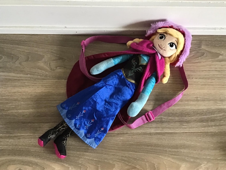 May include: A plush doll of a girl with long blonde hair wearing a blue dress, a pink cape, and a purple hood. The doll is lying on a wooden floor with a pink strap attached to the doll's back.