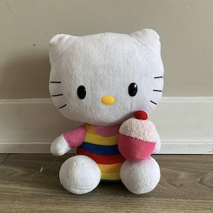 以下が含まれることがあります： 黒い目、ひげ、黄色い鼻を持つ白いぬいぐるみ猫。猫はカラフルなストライプのドレスを着て、赤いさくらんぼが付いたピンク色のカップケーキを持っています。おもちゃは木の床に座っています。