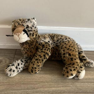 May include: A soft leopard toy with a tan and black spotted design. The stuffed animal has a white muzzle, tan nose, and black markings around the eyes. The toy is lying down on a wooden floor.