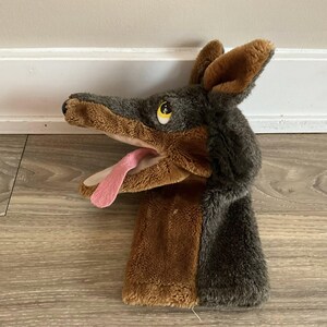 May include: A plush dog puppet with a gray and brown color scheme. The puppet has a long snout, floppy ears, and a pink tongue. The eyes are yellow. The puppet is made of soft, fuzzy material.