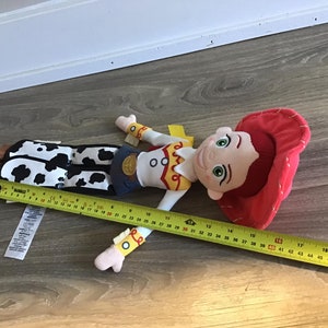 May include: A plush toy of Jessie from Toy Story. She is wearing her signature red cowboy hat, a yellow vest, and a black and white cow print skirt. She has brown hair and green eyes. The toy is approximately 40 inches long.