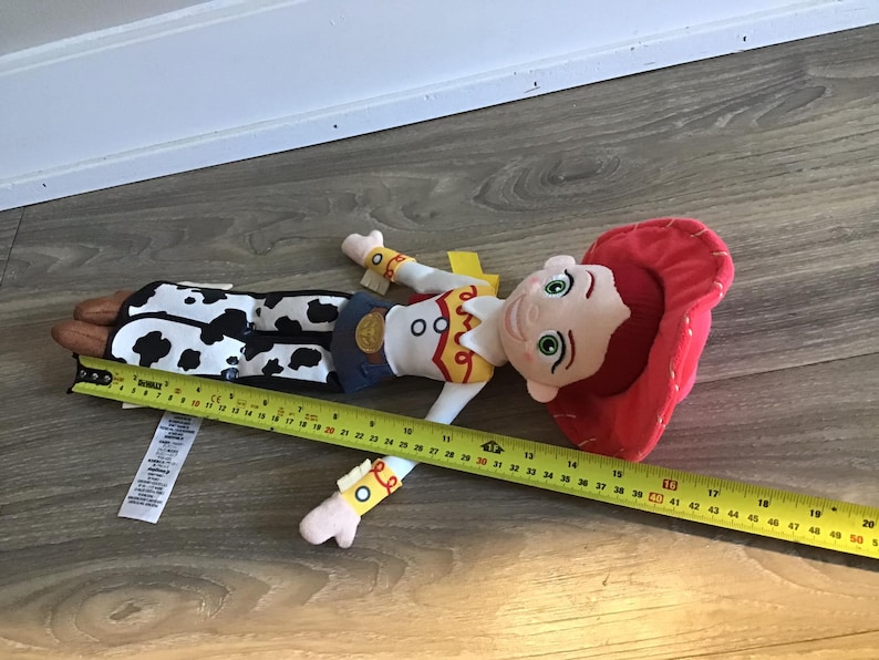May include: A plush toy of Jessie from Toy Story. She is wearing her signature red cowboy hat, a yellow vest, and a black and white cow print skirt. She has brown hair and green eyes. The toy is approximately 40 inches long.
