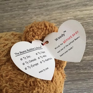 May include: A brown plush toy bear with a heart-shaped tag that reads "Woody the BEANIE BABY was one of the softest Beanic bears ever made!" The tag also includes the text "The Beanie Buddies Collection" and "www.ty.com".