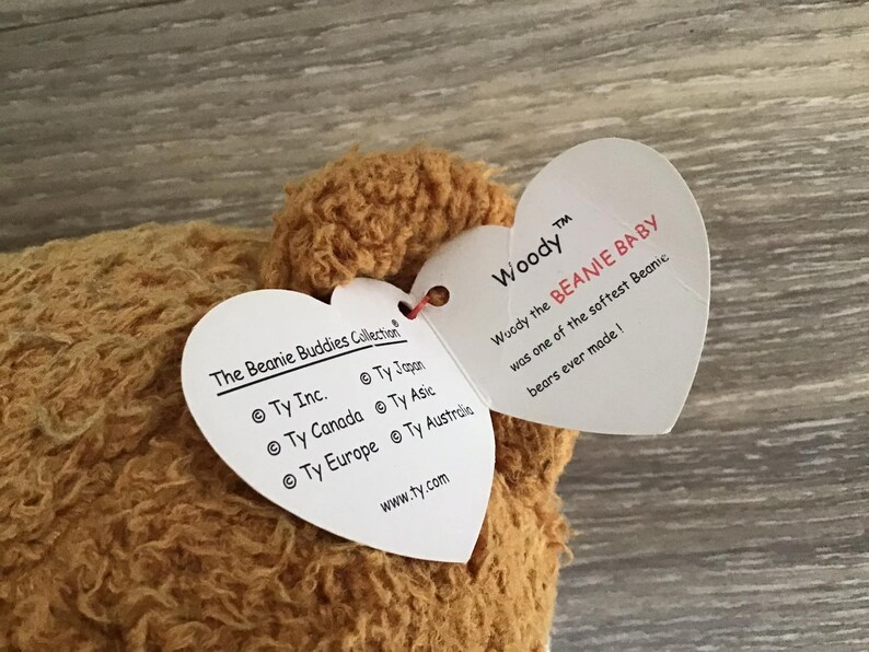 May include: A brown plush toy bear with a heart-shaped tag that reads "Woody the BEANIE BABY was one of the softest Beanic bears ever made!" The tag also includes the text "The Beanie Buddies Collection" and "www.ty.com".