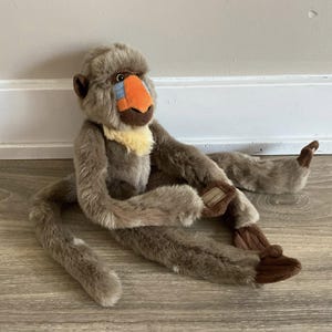 May include: A plush toy baboon with a brown body, long arms and legs, and a long tail. The baboon has a distinctive orange nose, a blue patch on its face, and a yellow collar. The toy is sitting on a wooden floor.