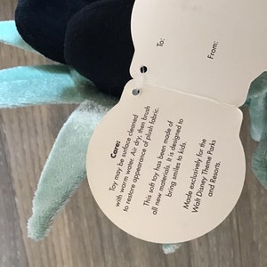 May include: A white tag with black text attached to a plush toy. The tag provides care instructions for the toy, stating that it can be surface cleaned with warm water and air dried. The tag also states that the toy is made of all new materials and is designed to bring smiles to kids. The tag also states that the toy is made exclusively for Walt Disney Theme Parks and Resorts.