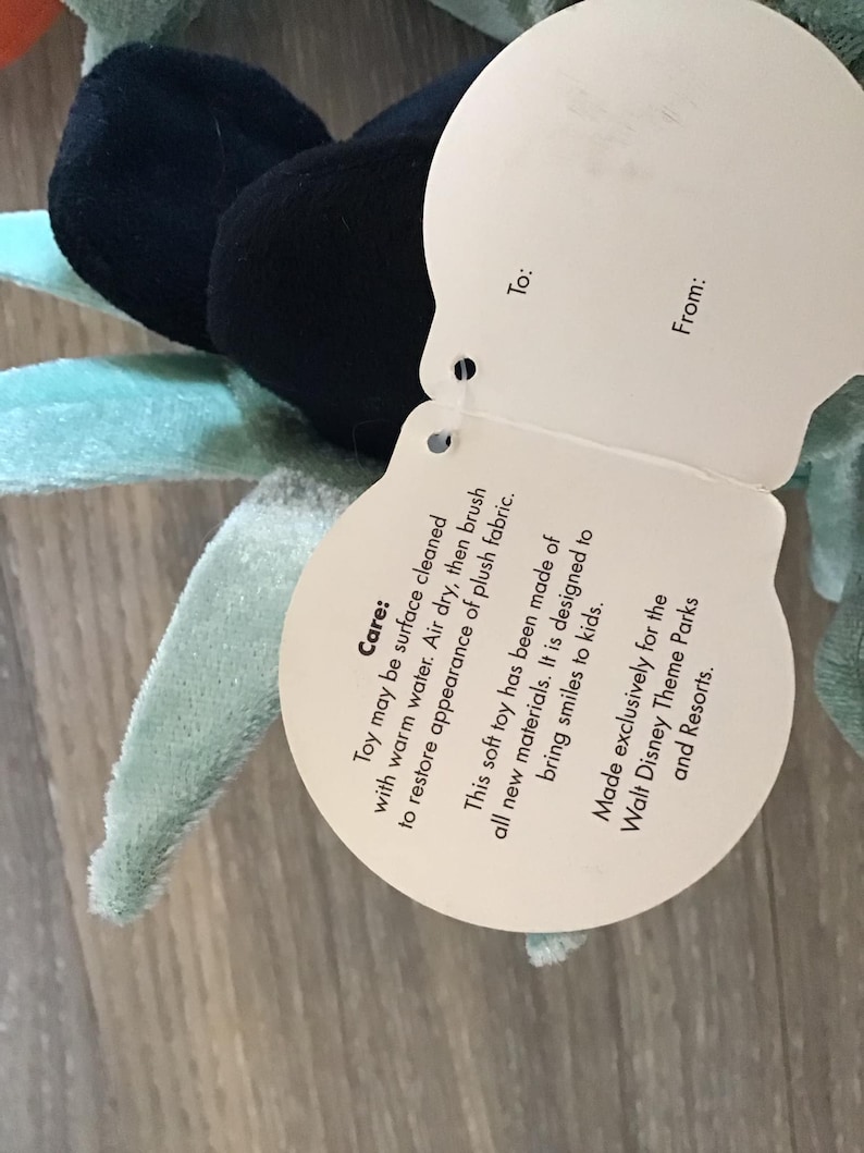 May include: A white tag with black text attached to a plush toy. The tag provides care instructions for the toy, stating that it can be surface cleaned with warm water and air dried. The tag also states that the toy is made of all new materials and is designed to bring smiles to kids. The tag also states that the toy is made exclusively for Walt Disney Theme Parks and Resorts.