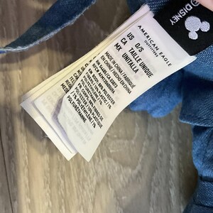 K&ouml;nnte beinhalten: Nahaufnahme eines wei&szlig;en Etiketts von American Eagle Outfitters mit dem Disney-Logo. Das Etikett hat schwarzen Text, der die Gr&ouml;&szlig;e als O/S und die Materialzusammensetzung angibt. Das Etikett ist an einem blauen Denim-Stoff befestigt.