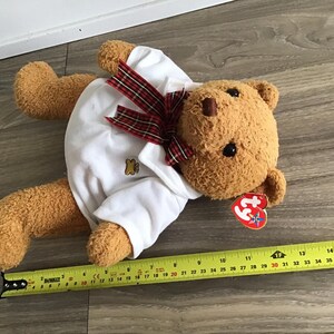May include: A brown teddy bear wearing a white polo shirt with a red and black plaid bow tie. The bear has a red Beanie Baby tag attached to its ear. The bear is lying on a wooden floor.