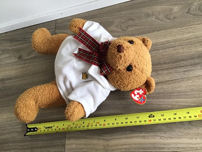May include: A brown teddy bear wearing a white polo shirt with a red and black plaid bow tie. The bear has a red Beanie Baby tag attached to its ear. The bear is lying on a wooden floor.