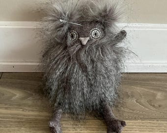 May include: A handmade gray owl plush toy with a fluffy body and head. The owl has patterned fabric eyes and feet, and a small beak. The owl is sitting on a wooden floor.