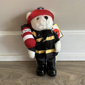 May include: A white teddy bear dressed as a New York City firefighter, wearing a black uniform with yellow stripes and a red helmet. The bear holds a red and white fire extinguisher. The uniform has "F.I." printed on it.