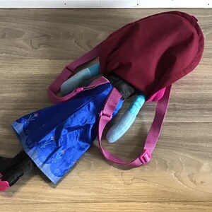 May include: A blue and purple backpack with a plush character attached. The character is wearing a blue dress with a floral pattern and has black boots. The backpack has a pink strap and a burgundy top.