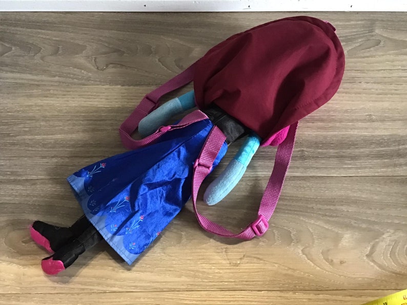 May include: A blue and purple backpack with a plush character attached. The character is wearing a blue dress with a floral pattern and has black boots. The backpack has a pink strap and a burgundy top.