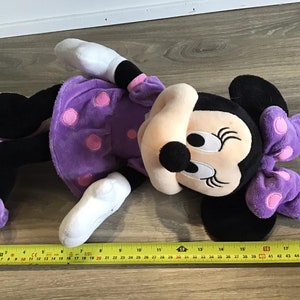 May include: A purple plush Minnie Mouse toy with pink polka dots and a purple bow. The toy is lying on a wooden floor with a measuring tape.