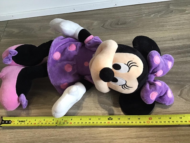 May include: A purple plush Minnie Mouse toy with pink polka dots and a purple bow. The toy is lying on a wooden floor with a measuring tape.