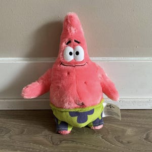 May include: A pink starfish plush toy with large white eyes, a small smile, and green and blue patterned shorts. The soft toy has a tag attached to its side.