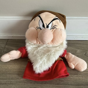 Puede incluir: Un títere de peluche de Gruñón de Blancanieves. El títere presenta un sombrero marrón, una barba blanca y una camisa roja. La cara de Gruñón está detallada con una nariz grande, cejas fruncidas y una expresión de mal humor.