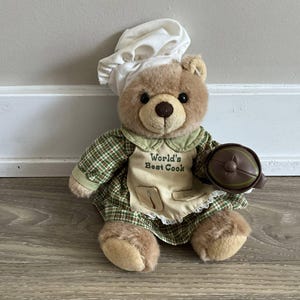 Może przedstawiać: Pluszowy miś w czapce kucharskiej i fartuchu. Na fartuchu napis "World's Best Cook". Miś ubrany jest w zieloną sukienkę w kratę i trzyma brązowy garnek. Miś siedzi na drewnianej podłodze.