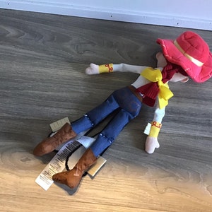 May include: A plush toy of Jessie from Toy Story. She is wearing a red hat, a yellow shirt, a blue denim skirt, and brown boots. She is lying on a wooden floor.