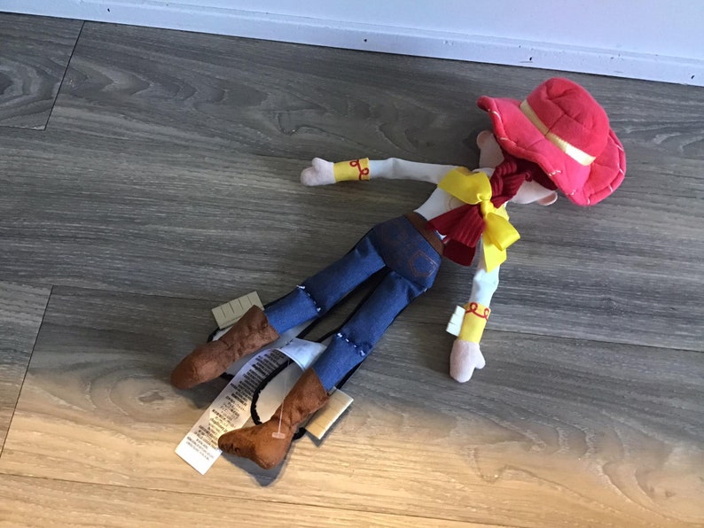 May include: A plush toy of Jessie from Toy Story. She is wearing a red hat, a yellow shirt, a blue denim skirt, and brown boots. She is lying on a wooden floor.