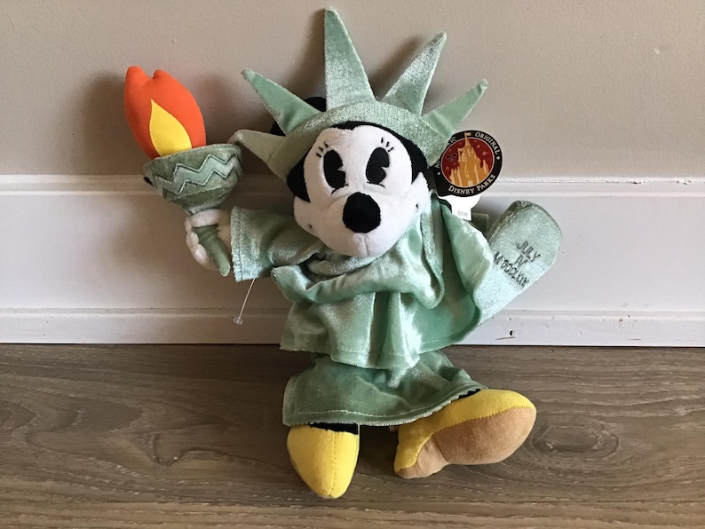 May include: A green plush Mickey Mouse dressed as the Statue of Liberty holding a torch. The plush is wearing a yellow hat and shoes. The tag says "July 4th in New York".