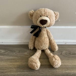 May include: A beige plush teddy bear with long arms and legs, wearing a black ribbon around its neck.
