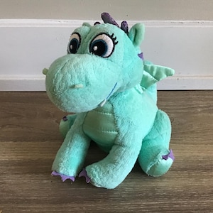 Puede incluir: Un dragón de peluche azul turquesa con detalles morados y grandes ojos negros. El dragón tiene una pequeña lengua rosa que sobresale y un cuerno morado en la cabeza.