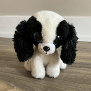 以下が含まれることがあります： 大きな青い目をした黒と白のぬいぐるみ犬。犬は垂れ耳と小さな黒い鼻をしています。
