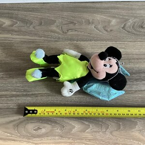 May include: A plush Mickey Mouse doll in a lime green outfit with blue accents. The doll has black ears, a pink face, and white gloves. The doll is lying on a blue pillow. A measuring tape is next to the doll.