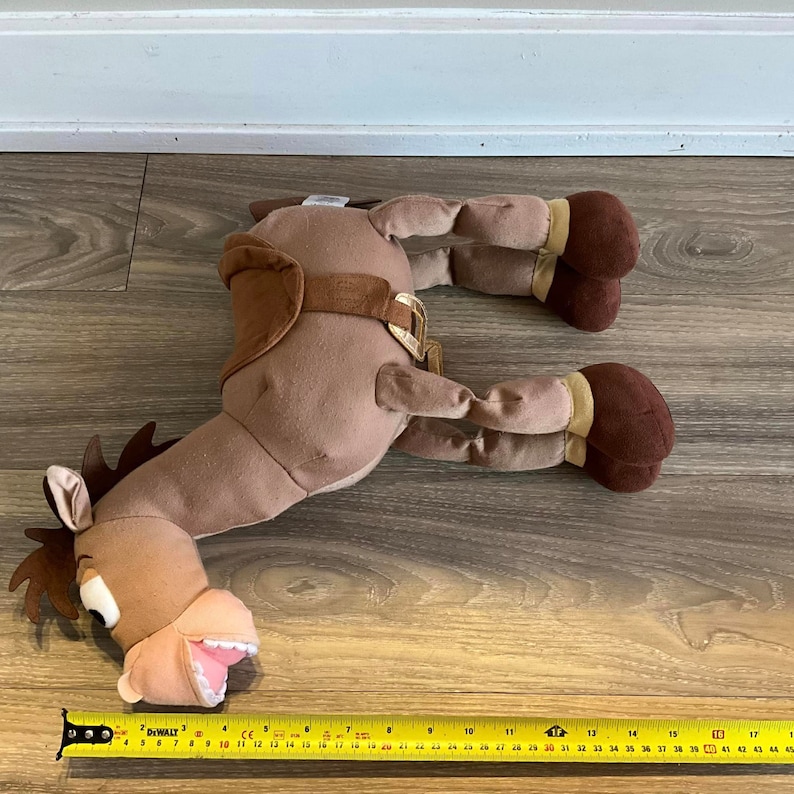 Puede incluir: Caballo de peluche marr&oacute;n con una silla de montar y una amplia sonrisa. El caballo est&aacute; acostado en un suelo de madera con una cinta m&eacute;trica en primer plano.