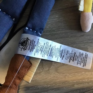 May include: A blue denim and brown plush toy with a white tag that reads "Disney Made in Vietnam Fabriqua Vietnam Hecho en Vietnam" and includes additional information about the product and the company.