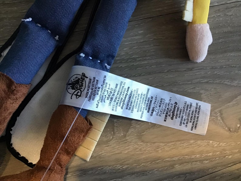 May include: A blue denim and brown plush toy with a white tag that reads "Disney Made in Vietnam Fabriqua Vietnam Hecho en Vietnam" and includes additional information about the product and the company.