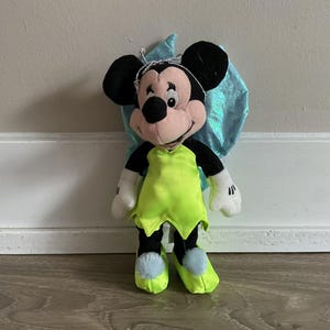 May include: A plush Minnie Mouse doll dressed as a fairy. The doll has black ears, a pink face, and is wearing a lime green dress and shoes. It has blue wings and a silver tiara. The doll's arms and hands are white.