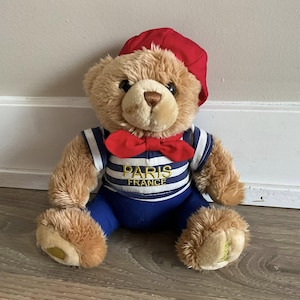 May include: A plush teddy bear wearing a red beret and bow tie. The bear is dressed in a blue and white striped shirt and blue pants. The shirt has the words "PARIS FRANCE" embroidered on it.
